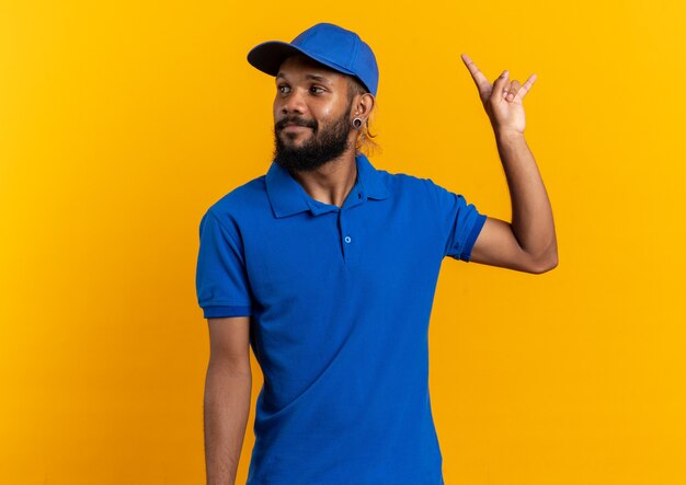 pleased young delivery man gesturing horns and looking at side isolated on orange wall with copy space