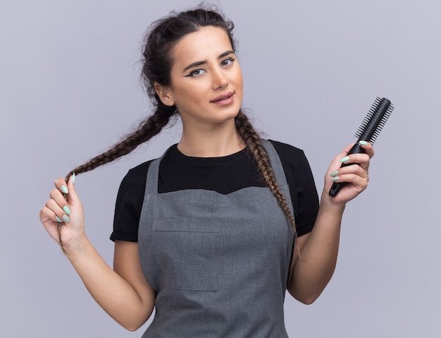 Pleased young female barber in uniform holding comb and grabbed hair isolated on white wall