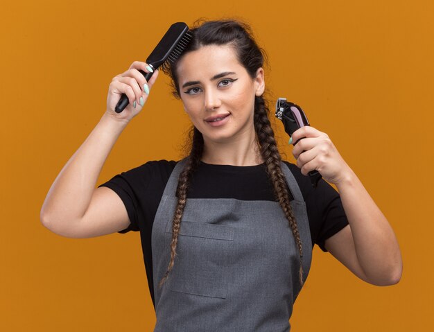 Pleased young female barber in uniform holding hair clippers and combing hair isolated on orange wall