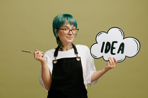 Pleased young female barber wearing glasses uniform holding scissors and idea bubble looking at camera isolated on olive green background