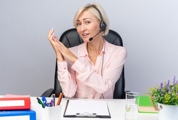 Pleased young female call center operator wearing headset sitting at table with office tools holding hands together isolated on white wall