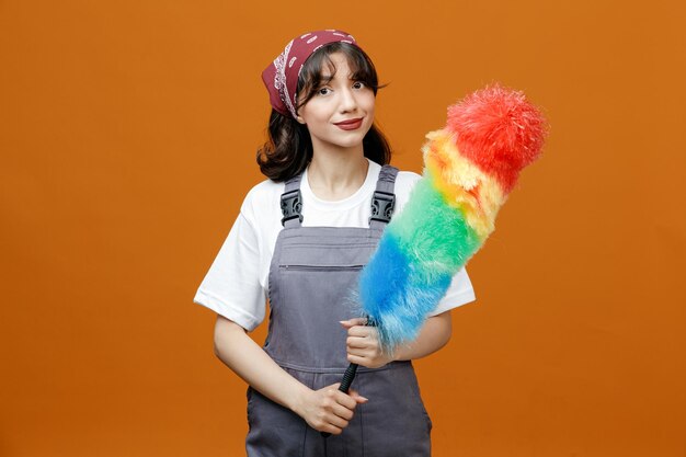 Pleased young female cleaner wearing uniform and bandana holding feather duster with both hands looking at camera isolated on orange background