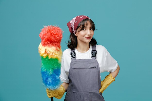 Pleased young female cleaner wearing uniform bandana and rubber gloves holding feather duster looking at camera while keeping hand on waist isolated on blue background