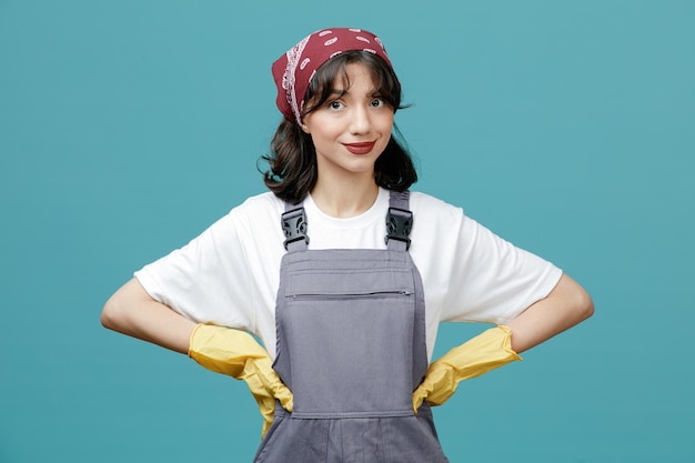 Pleased young female cleaner wearing uniform bandana and rubber gloves keeping hands on waist looking at camera isolated on blue background