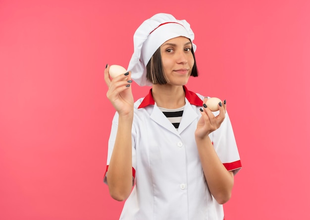 Pleased young female cook in chef uniform holding eggs isolated on pink wall