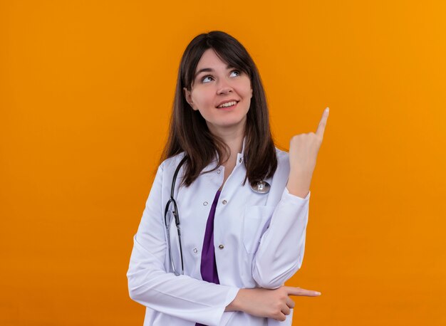 Pleased young female doctor in medical robe with stethoscope points up and looks up on isolated orange background with copy space