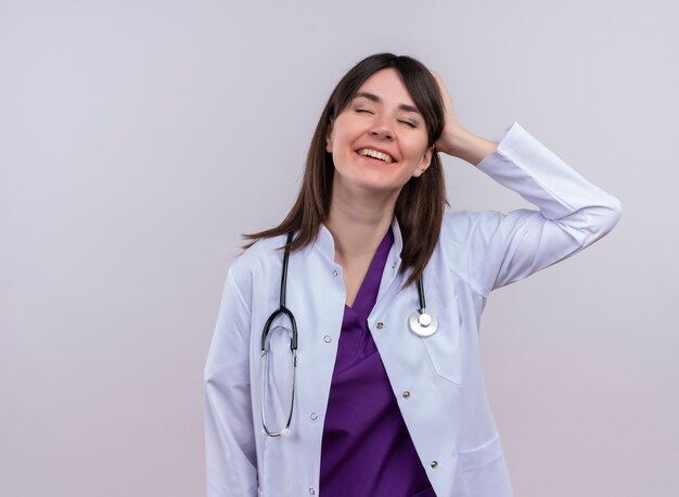 Pleased young female doctor in medical robe with stethoscope puts hand behind head on isolated white background with copy space