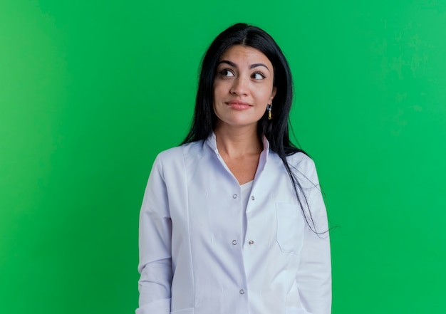 Free photo pleased young female doctor wearing medical robe looking at side isolated on green wall with copy space