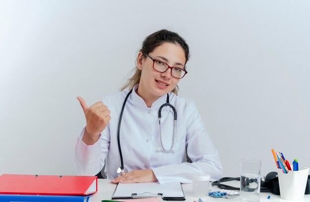 Pleased young female doctor wearing medical robe and stethoscope and glasses sitting at desk with medical tools looking showing thumb up isolated