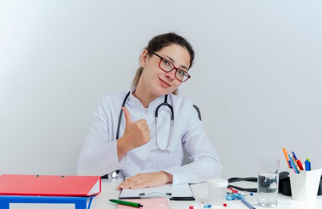 Pleased young female doctor wearing medical robe and stethoscope and glasses sitting at desk with medical tools looking showing thumb up isolated