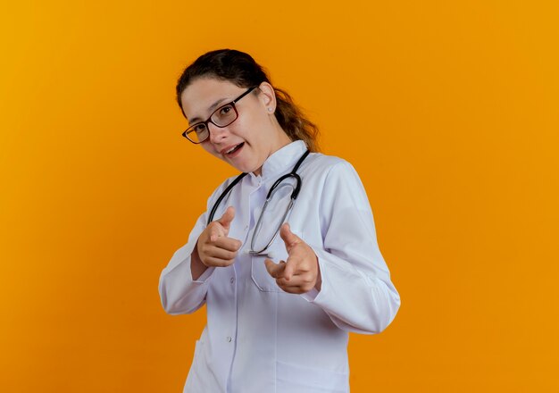 Pleased young female doctor wearing medical robe and stethoscope with glasses showing you gesture isolated