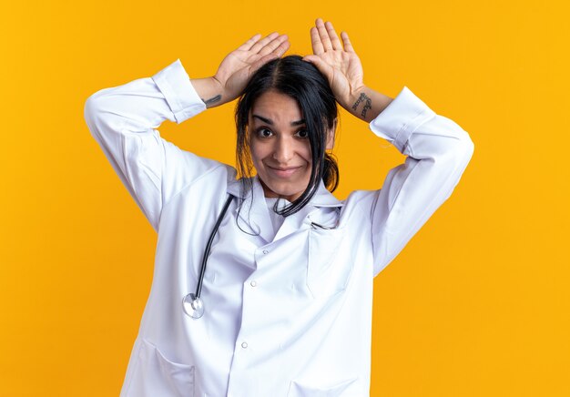 Pleased young female doctor wearing medical robe with stethoscope showing bunny ears gesture isolated on yellow wall