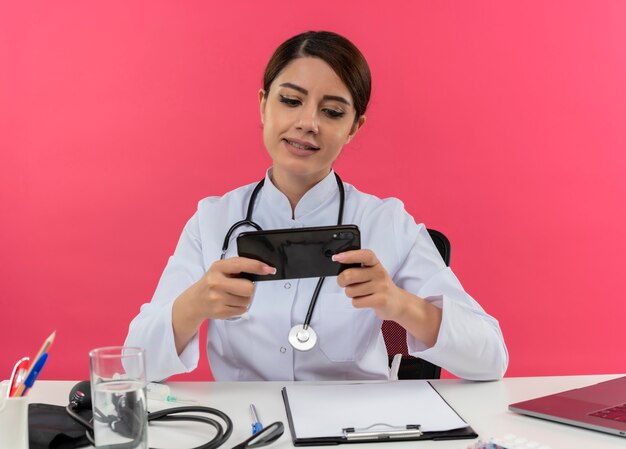 Pleased young female doctor wearing medical robe with stethoscope sitting at desk work on computer with medical tools playing game on phone with copy space