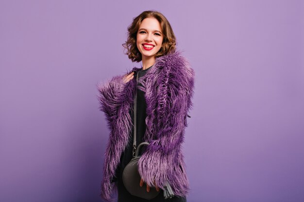 Pleased young female model with sincere smile posing on purple background, holding gray purse