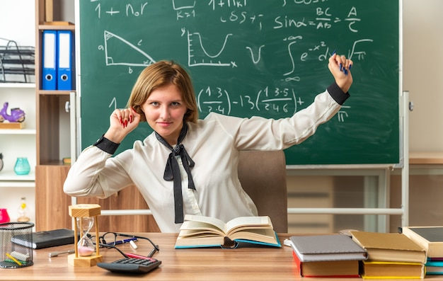 Free photo pleased young female teacher sits at table with school supplies in classroom