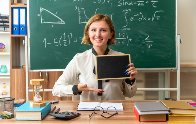 Pleased young female teacher sits at table with school tools holding and points with hand at mini blackboard in classroom