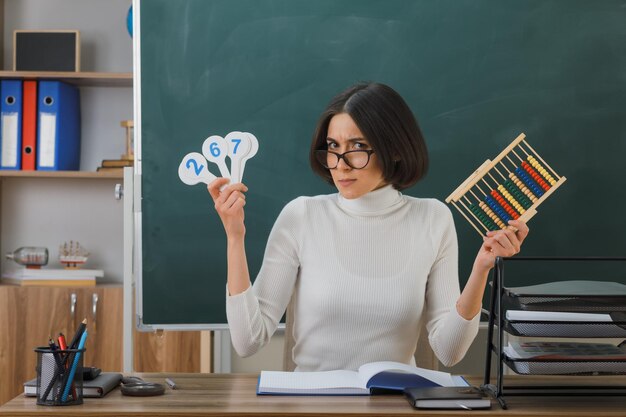 pleased young female teacher wearing glasses holding abacus with number fan sitting at desk with school tools on in classroom