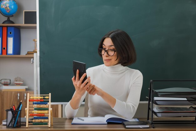 pleased young female teacher wearing glasses holding and looking at calculator sitting at desk with school tools on in classroom