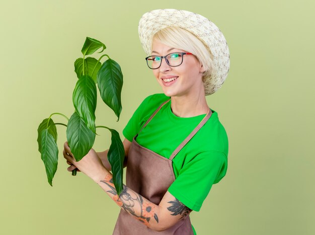 Pleased young gardener woman with short hair in apron and hat holding plant lookign at camera with smile on face standing over light background