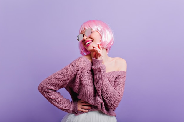 Pleased young girl in periwig and sunglasses having fun  Photo of wonderful female model with pink hair smiling while dancing on purple wall.