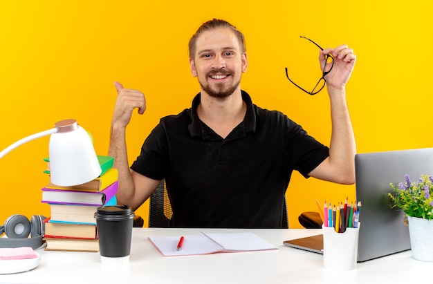 Pleased young guy student sitting at table with school tools holding glasses showing thumb up isolated on orange wall