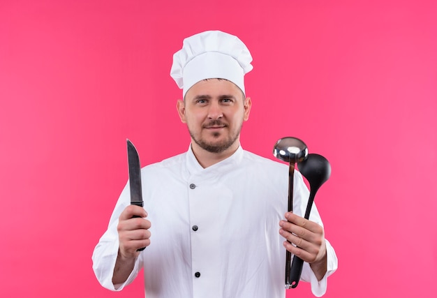 Free photo pleased young handsome cook in chef uniform holding knife and ladles isolated on pink space