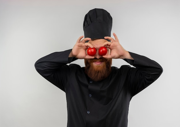 Free photo pleased young handsome cook in chef uniform putting tomatoes on eyes isolated on white space