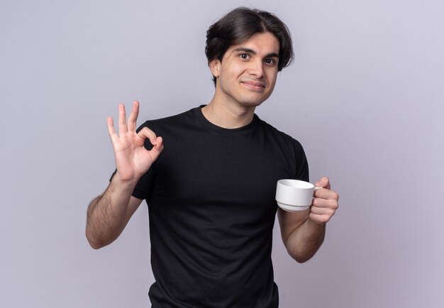 Free Photo pleased young handsome guy wearing black t-shirt holding cup of coffee showing okay gesture isolated on white wall