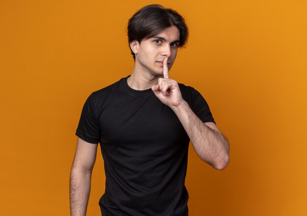 Pleased young handsome guy wearing black t-shirt showing silence gesture isolated on orange wall with copy space