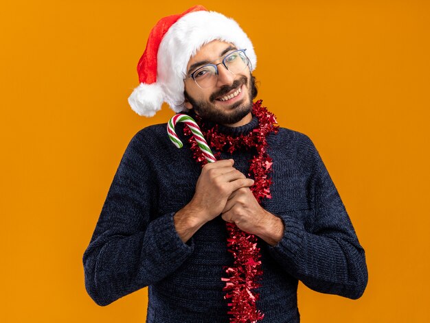 pleased young handsome guy wearing christmas hat with garland on neck holding christmas candy isolated on orange wall