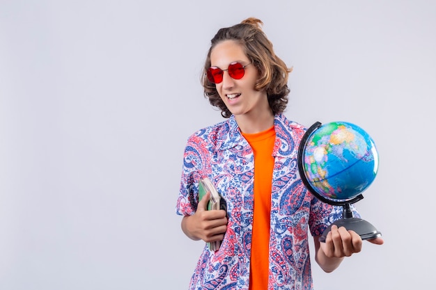 Free Photo pleased young handsome guy wearing red sunglasses holding globe and notebook looking at camera with confident smile standing over white background