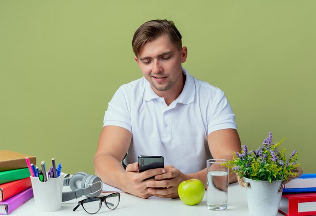 Pleased young handsome male student sitting at desk with school tools holding and looking at phone isolated on olive green