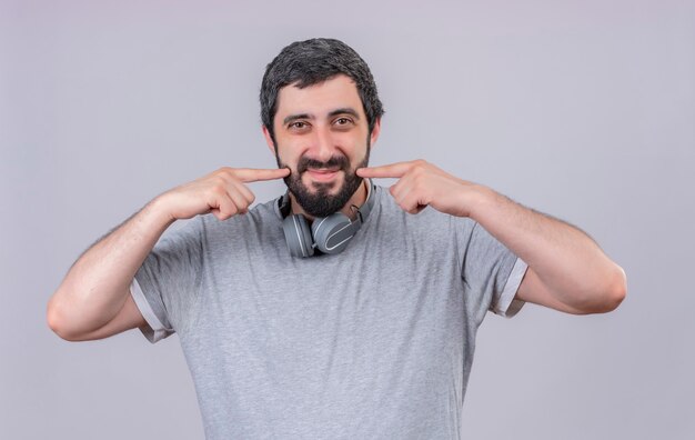 Pleased young handsome man wearing headphones on neck and pointing at his smile isolated on white wall