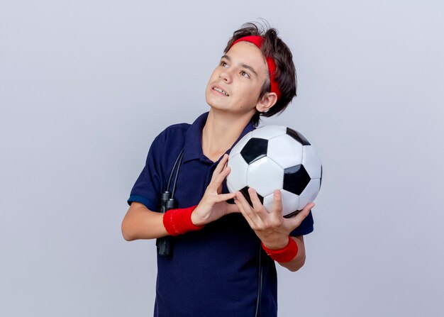 Pleased young handsome sporty boy wearing headband and wristbands with dental braces and jump rope around neck holding soccer ball looking up isolated on white background with copy space