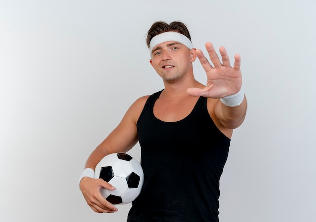 Pleased young handsome sporty man wearing headband and wristbands holding soccer ball and stretching out hand towards front isolated on white wall