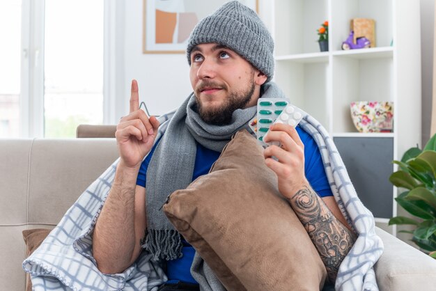 pleased young ill man wearing scarf and winter hat wrapped in blanket sitting on sofa in living room holding pillow holding packs of pills looking and pointing up