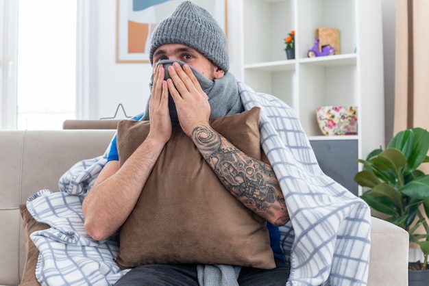 pleased young ill man wearing scarf and winter hat wrapped in blanket sitting on sofa in living room holding pillow looking at front covering mouth with scarf