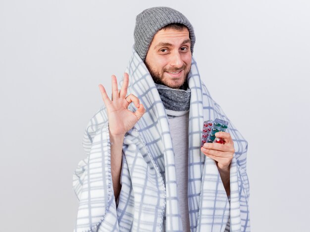 Pleased young ill man wearing winter hat and scarf wrapped in plaid holding pills