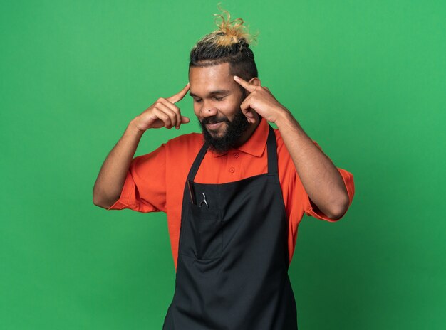 Pleased young male barber wearing uniform doing think gesture with closed eyes isolated on green wall