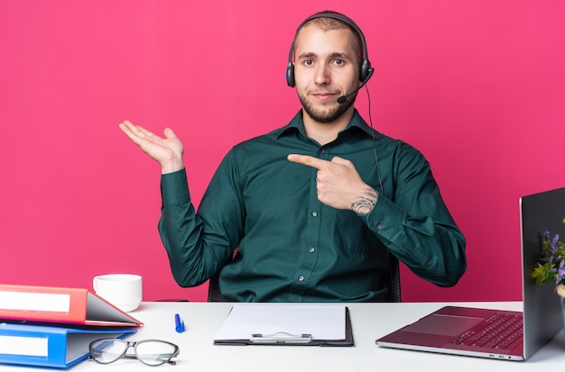 Free photo pleased young male call center operator wearing headset sitting at desk with office tools pretending holding and points at something