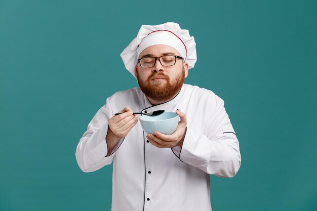 Pleased young male chef wearing glasses uniform and cap holding spoon and empty bowl pretend smelling aroma of food with closed eyes isolated on blue background