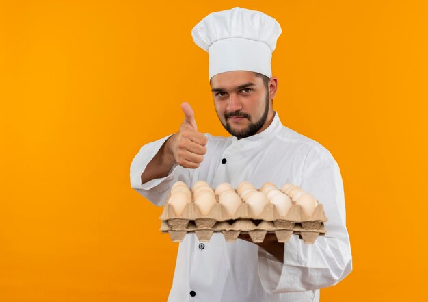 Pleased young male cook in chef uniform holding carton of eggs and showing thumb up isolated on orange space 