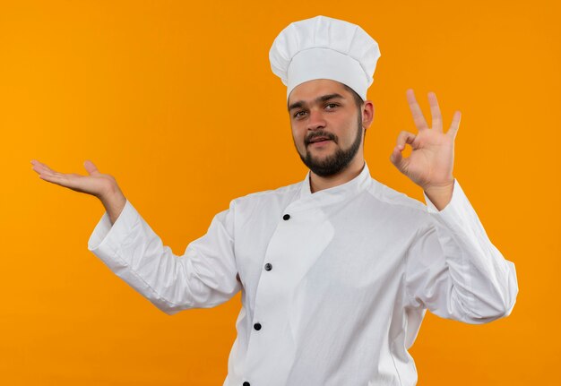 Pleased young male cook in chef uniform showing empty hand and doing ok sign isolated on orange space