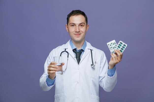 Pleased young male doctor wearing medical robe and stethoscope around neck looking at camera holding glass of water showing packs of pills isolated on purple background