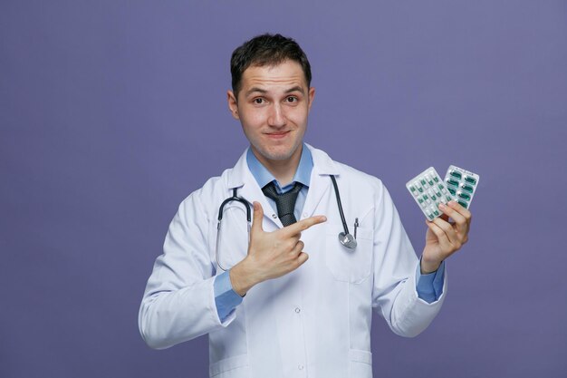 Pleased young male doctor wearing medical robe and stethoscope around neck looking at camera showing packs of pills pointing at them isolated on purple background