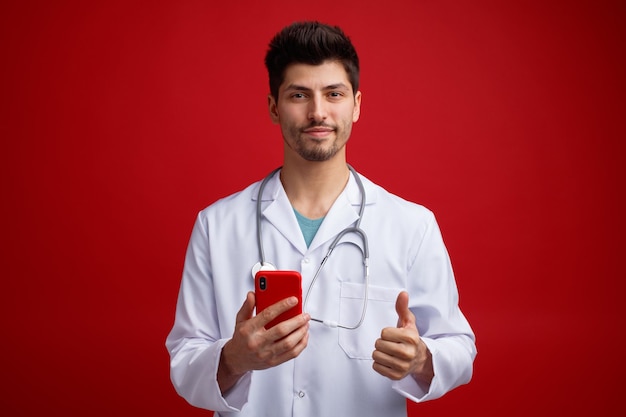 Pleased young male doctor wearing medical uniform and stethoscope around his neck holding mobile phone looking at camera showing thumb up isolated on red background