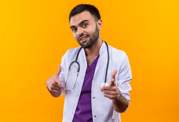 Pleased young male doctor wearing stethoscope medical gown showing you gesture on isolated yellow background