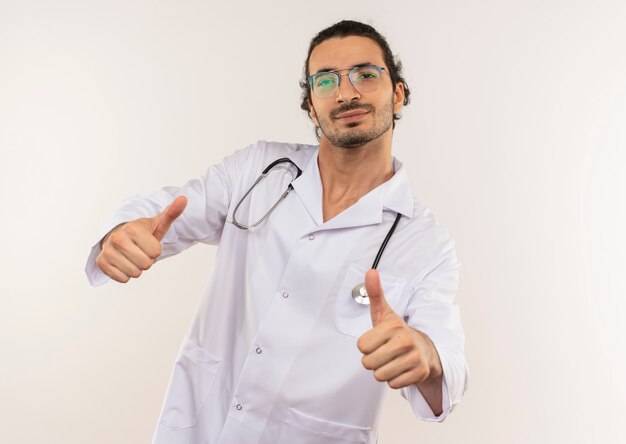 Pleased young male doctor with optical glasses wearing white robe with stethoscope his thumbs up on isolated white wall with copy space