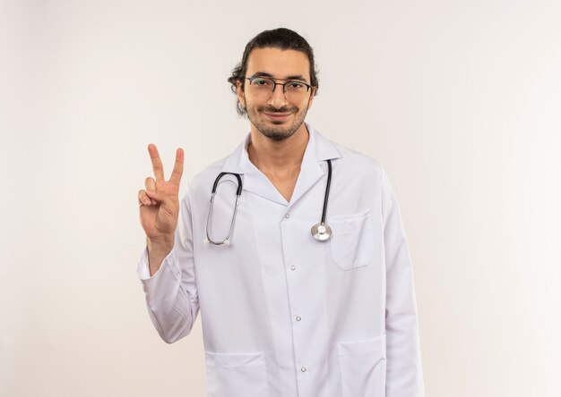 Pleased young male doctor with optical glasses wearing white robe with stethoscope showing peace gesture on isolated white wall with copy space