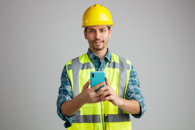 Pleased young male engineer wearing safety helmet and uniform holding mobile phone with both hands looking at camera isolated on white background